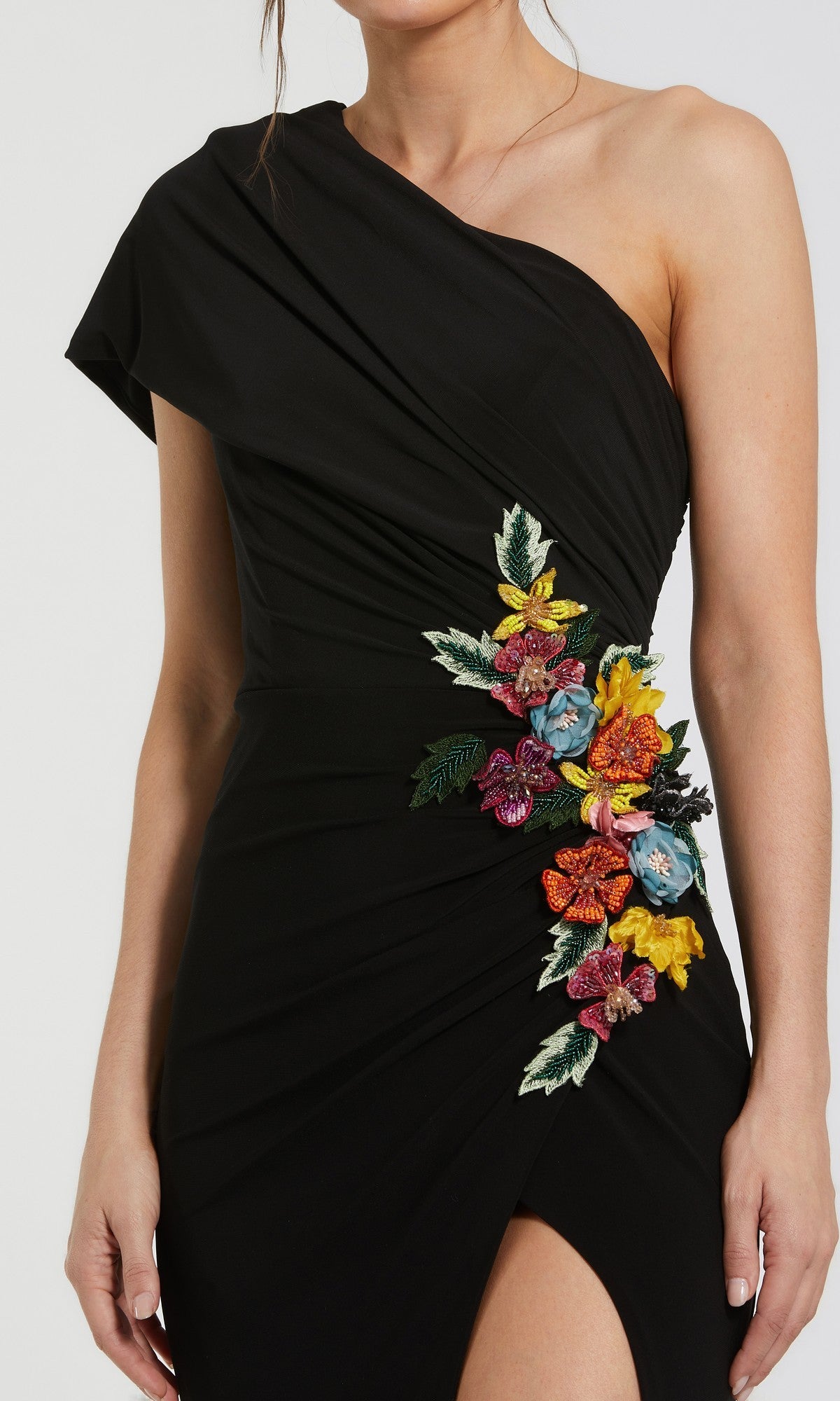 A woman wearing the Mac Duggal 11769 long formal dress—an elegant black, one-shoulder jersey gown beautifully embellished with colorful embroidered flowers and eye-catching 3D floral appliqué along the side, finished with a bold thigh-high slit.
